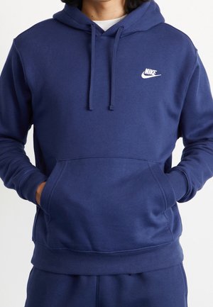 Osoba ubrana w granatową bluzę z kapturem Nike z rękami w przedniej kieszeni, z białym logo Nike na lewej piersi.