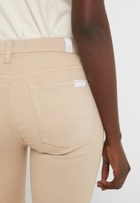 Pantalones color beige con un diseño ajustado, que cuentan con dos bolsillos traseros y detalles de costura sutiles. La tela es suave y tiene un ligero brillo.