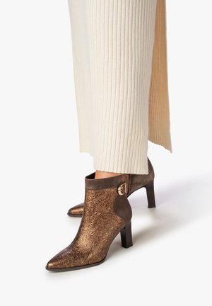 Bottines en bronze avec une finition texturée, bout pointu et un accent de boucle dorée sur un panneau marron ; talon carré. Portées avec un pantalon crème côtelé.