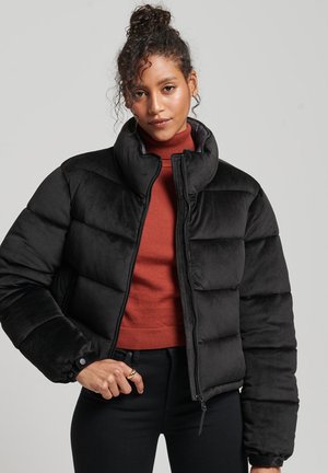 Frau mit lockigem Haar, das zu einem Dutt frisiert ist, trägt eine schwarze Daunenjacke über einem rostfarbenen Rollkragenpullover und posiert mit einer Hand an der Taille vor einem grauen Hintergrund.