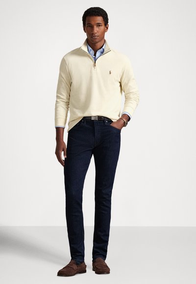 Muž oblečený v krémovém svetru s čtvrtým zipem přes modrou košili, tmavých úzkých džínách, hnědém koženém opasku a hnědých semišových mokasínách stojí s jednou rukou v kapse.