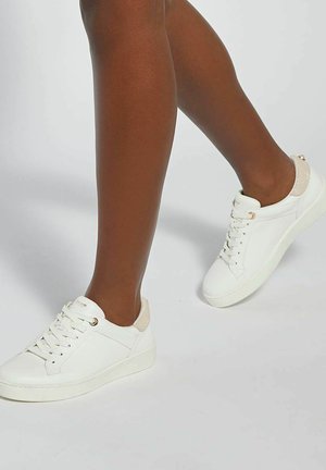 Jambes portant des baskets blanches à lacets avec des accents beige au talon, positionnées comme si elles marchaient sur un fond clair uni.