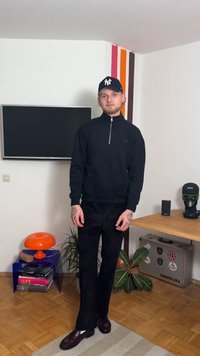 Μαύρο πουλόβερ με φερμουάρ, μαύρο παντελόνι και σκούρα παπούτσια. Στέκεται σε ένα δωμάτιο με πορτοκαλί φωτιστικό, φυτά και μια τηλεόραση στο φόντο.