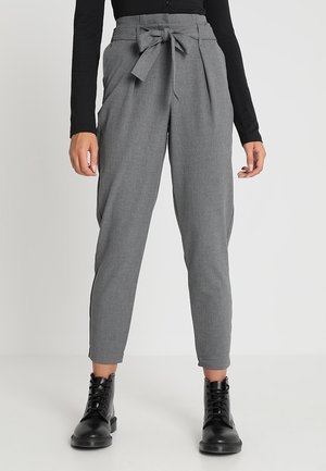 Vrouw draagt een grijze, hooggetailleerde, taps toelopende broek met een strik aan de voorkant, gecombineerd met zwarte veteren enkellaarsjes en een zwarte top met lange mouwen.