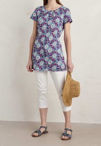 Túnica azul de flores con mangas cortas, que presenta patrones blancos y morados. Combinada con capris blancos y sandalias grises, sosteniendo un sombrero de paja.