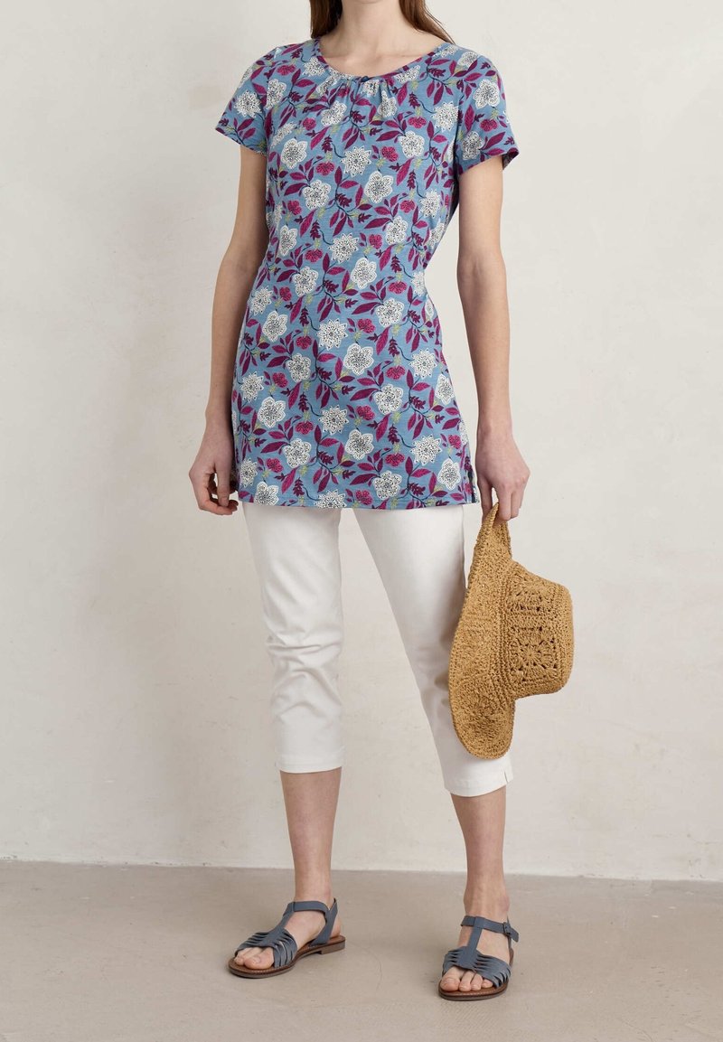 Túnica azul de flores con mangas cortas, que presenta patrones blancos y morados. Combinada con capris blancos y sandalias grises, sosteniendo un sombrero de paja.