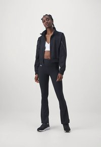 Sort cropped jakke med stående krave og elastiske manchetter, kombineret med sorte vide leggings og sorte sportssko, båret ovenpå en hvid top.