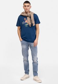 Camiseta azul marino con estampado gráfico colorido y texto, combinada con jeans azul claro y zapatos beige. Un suéter color beige está colocado sobre los hombros.