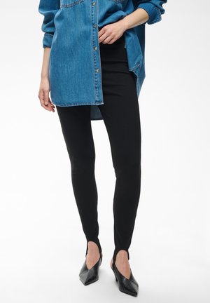 Personne portant un pantalon noir ajusté avec des étriers, des talons pointus noirs et une chemise en jean bleue oversize aux manches retroussées.