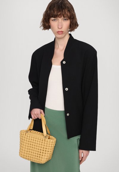 Zwarte cropped jasje met een ronde halslijn en zilveren knopen, gecombineerd met een witte top en groene rok. Houdt een kleine gele handtas van gevlochten materiaal vast.