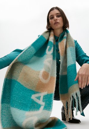 Femme aux cheveux courts et foncés agenouillée, portant une chemise bleu sarcelle et des bottes noires, tenant une grande écharpe bleue, beige et blanche à motifs avec des franges.
