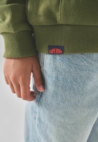 Olivově zelená mikina s ribovanými manžetami, s modro-oranžovým štítkem "Superdry", doplněná o světle modré džíny, se zaměřením na texturu.