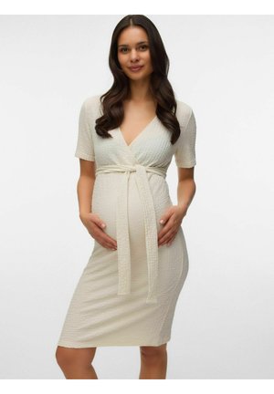 Femme enceinte aux longs cheveux foncés portant une robe portefeuille crème texturée, tenant son ventre et souriant sur un fond uni.