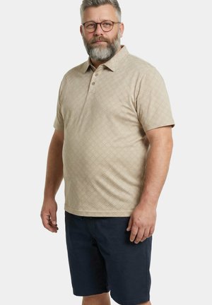 Uomo di mezza età con barba grigia e occhiali, indossa una polo beige a fantasia e pantaloncini blu navy, in piedi davanti a uno sfondo semplice.