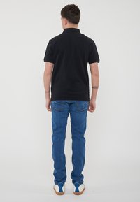 Polo shirt nero a maniche corte con colletto, abbinato a jeans blu slim-fit e sneakers bianche con dettagli blu. Sfondo bianco uniforme.