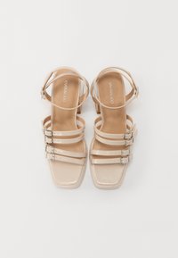 Sandales beige clair avec un orteil carré et une finition brillante. Dotées de trois fines sangles avec des boucles en argent, ainsi qu'une sangle de cheville.