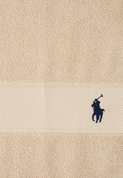 Πετσέτα μπεζ με υφή, διακοσμημένη με υφασμένη λωρίδα που έχει κεντημένη μια φιγούρα παίκτη πόλο με μπλε ναυτικό ιππέα κοντά στην άκρη.