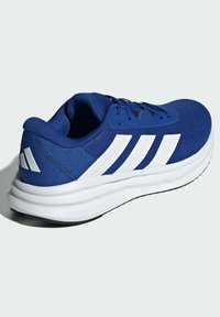 Modrá sportovní bota s textilem s texturou, bílým designem se třemi pruhy a silnou bílou gumovou podrážkou; má polstrovaný límec a jazyk.