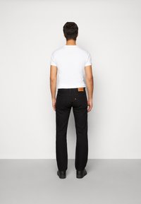 Jeans negros con corte ajustado, que presentan un parche de cuero marrón y una etiqueta de logo roja en la parte trasera. Combinados con una camiseta blanca básica y zapatos negros.