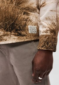 Καφέ patterned μακρυμάνικο πουκάμισο με ετικέτα υφάσματος, σε συνδυασμό με γκρι κολάν από κωδούρο. Το πουκάμισο διαθέτει ένα σχέδιο εμπνευσμένο από τη φύση.