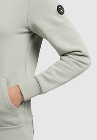 Braccio sinistro di una persona che indossa una felpa grigia chiara, con la mano nella tasca a marsupio e una toppa nera sulla manica superiore.