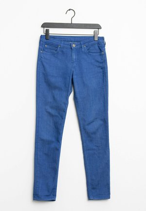Pantalones vaqueros de denim azul colgados en una percha negra. Presentan un corte ajustado, diseño de cinco bolsillos y pequeños detalles en botón y remaches de metal.