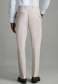 Pantaloni bej, croiți cu o finisare netedă, având două buzunare la spate și o croială slim, asortați cu pantofi închiși la culoare.
