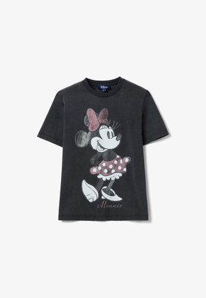 T-shirt noir à manches courtes présentant Minnie Mouse de style vintage, portant une jupe et un nœud rose à pois, debout les mains sur les hanches.