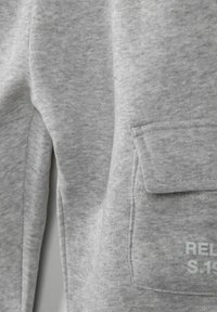 Grå sweatshirttyg med fin textur, som har en ficka på sidan och tryckt text i ljusgrå, som anger märke och stilinformation.