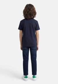 Παιδί με σγουρά καστανά μαλλιά, φοράει μπλε μπλουζάκι ναυτικού χρώματος, σκουρόχρωμα μπλε τζιν και λευκά αθλητικά παπούτσια με πράσινες λεπτομέρειες, στέκεται γυρισμένο με την πλάτη.