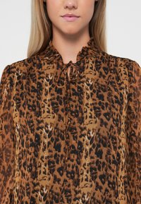 Bluzica s smeškom leopard uzorkom, s volanom na izrezu i mašnom sprijeda. Prozirna tkanina s laganom teksturom i dugim rukavima.