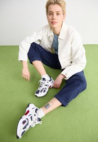 Vita sneakers med marinblå och röda accenter; mesh och syntetiska material; i kombination med marinblå capribyxor och en ljus jacka.