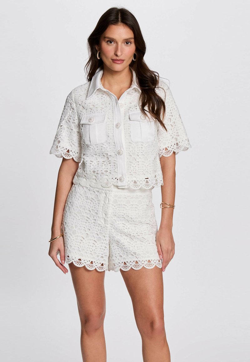 Completo in due pezzi bianco in crochet, composto da una camicia a maniche corte con bottoni e due tasche sul petto, abbinata a pantaloni corti con bordi scallop.