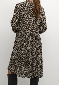 Femme portant une robe genou à fleurs sombres avec des manches longues, associée à des bottes noires jusqu'aux genoux, se tenant devant un fond uni.