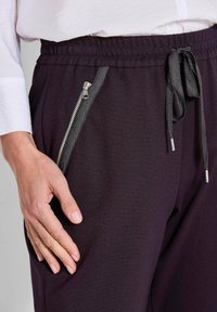 Zwarte joggerbroek van zacht materiaal, met een brede tailleband, zijzakken met rits en zilverkleurige hardware, en een trekkoorddetail.