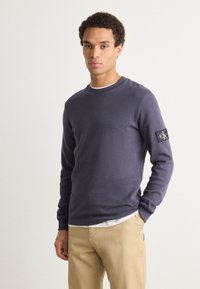 Tengerészeti crewneck pulóver texturált anyagból, logóvalsókkal az ujjon. Világos színű nadrággal párosítva.