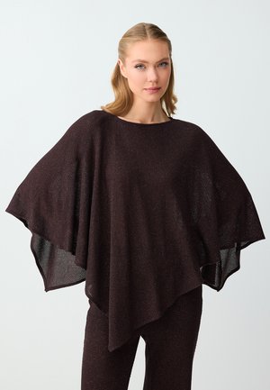 Femme aux cheveux clairs portant un poncho en maille marron foncé ample à larges manches, associé à un pantalon droit assorti, debout devant un fond gris clair.