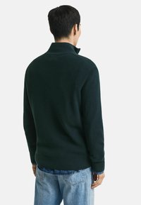 Man in een donkergroene ribbeltrui met hoge kraag, met lichte spijkerbroek, van achteren kijkend tegen een witte achtergrond.
