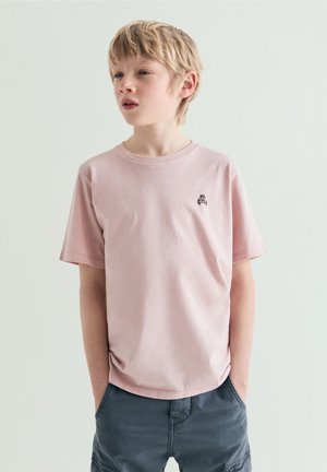 Blond chłopiec w jasnoróżowej koszulce z małym logo czaszki i ciemnoszarych krótkich spodenkach, stoi z rękami w kieszeniach, patrząc w lewo.