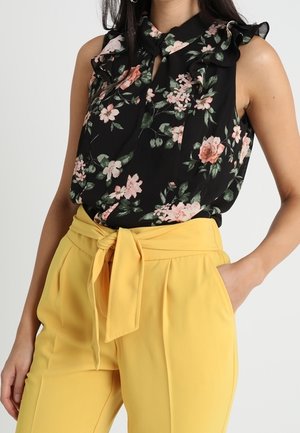 Kobieta ubrana w czarną bezrękawnikową bluzkę w kwiaty z falbanami oraz żółte spodnie z wysokim stanem, związane z przodu kokardą.
