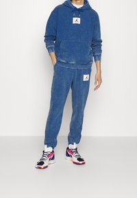 Blaues Acid-Wash-Hoodie mit Kängurutasche und Logopatch, kombiniert mit passenden Jogginghosen mit Logopatch. Bunte Sportschuhe vervollständigen das Outfit.