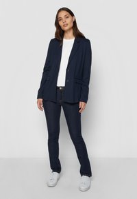 Marineblazer med to knapper, hakkeslag og sidelommer; kombinert med en hvit t-skjorte og mørke denimjeans. Hvite sneakers fullfører antrekket.