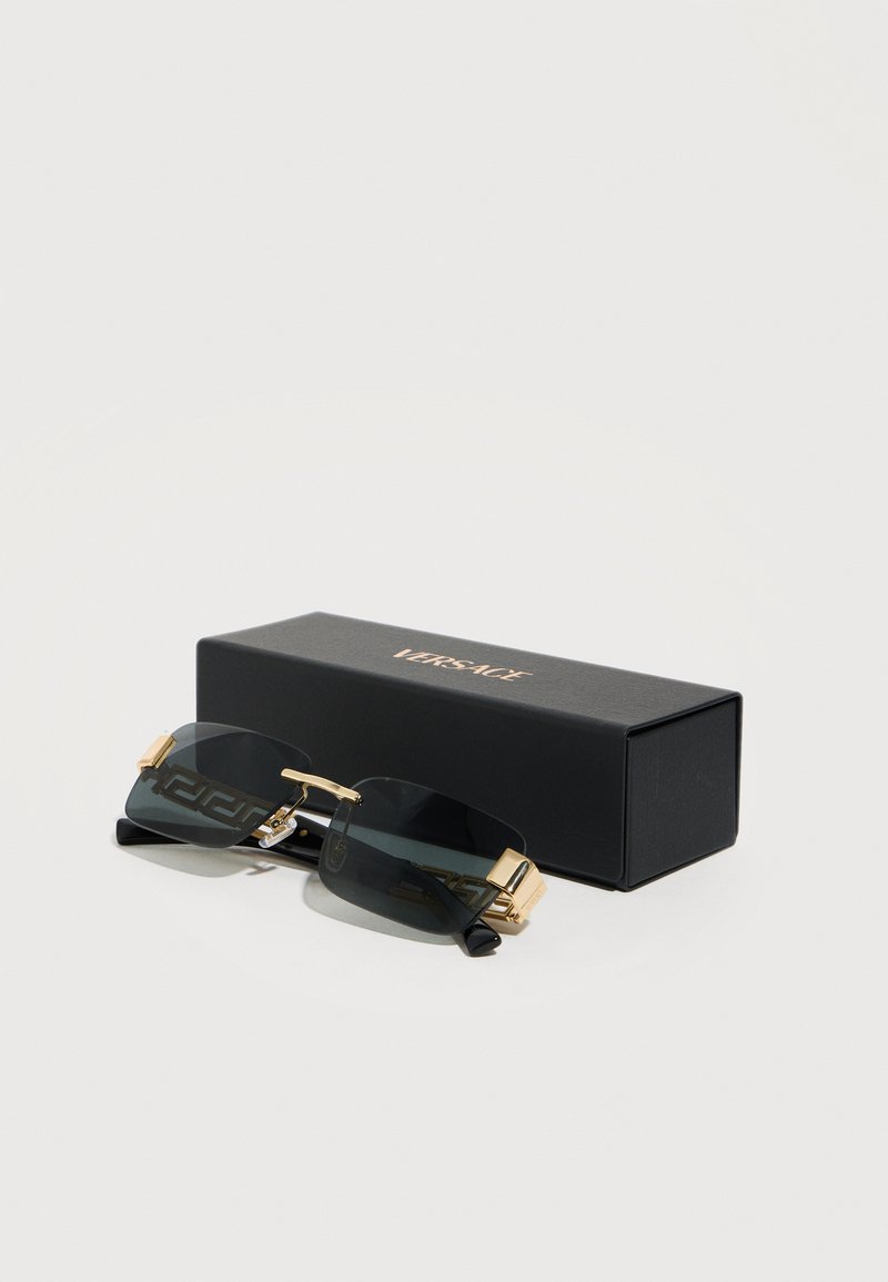 Rimless dark sunglasses with gold accents placed next to a black rectangular Versace case on a light surface.