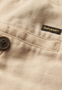 Tela beige con un tejido texturizado, que presenta un botón oscuro y una etiqueta negra con "SUPERDRY" en letras doradas, posicionada cerca de una costura.