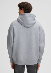 Un uomo di spalle indossa una felpa grigia chiara e semplice con il cappuccio abbassato e jeans grigi scuri, davanti a uno sfondo chiaro e uniforme.