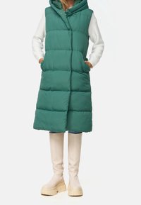 Gilet verde imbottito con cappuccio, caratterizzato da un design a righe orizzontali e tasche frontali, abbinato a stivali bianchi fino al ginocchio e jeans.