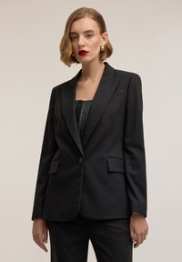 Zwarte op maat gemaakte blazer met een enkele knoopsluiting, ingezette revers en twee voorkanten zakken, vervaardigd uit een gladde stof.