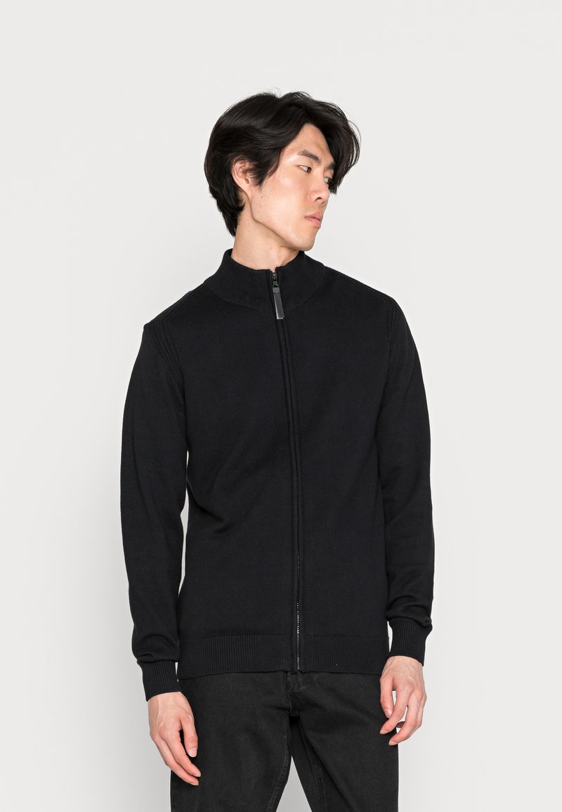 Svart zip-up-tröja med hög krage, ribbad textur och långa ärmar. Har en framdragkedja och åtsittande manschetter i nederkant.