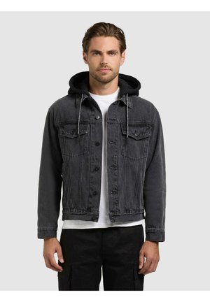 Mann iført svart denimjakke med påsatt svart hette, hvit t-skjorte og svarte bukser, stående mot en ensfarget grå bakgrunn.