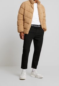 Beige polyester puffjacka med quiltat mönster, ovanpå en vit T-shirt. Svarta joggingbyxor med en märkeskant i elastiskt material och vita sneakers.